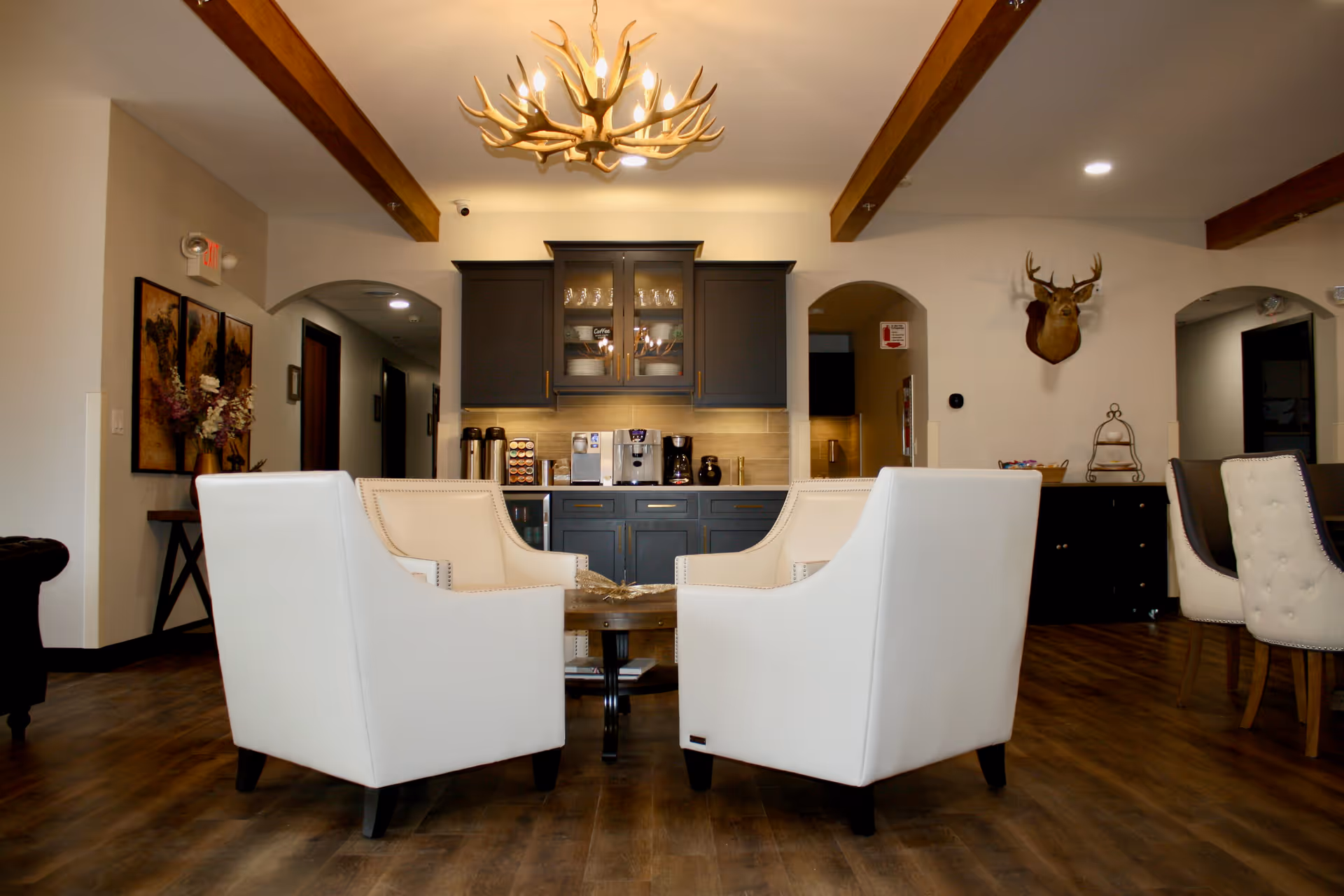 A cozy common area with four white armchairs arranged around a wooden coffee table. Behind the seating area is a dark gray cabinet with glass doors displaying dishes and coffee supplies. The room features wooden ceiling beams, a chandelier made of antlers, wooden flooring, and a mounted deer head on the wall. There are archways leading to other rooms and decorative artwork on the walls.
