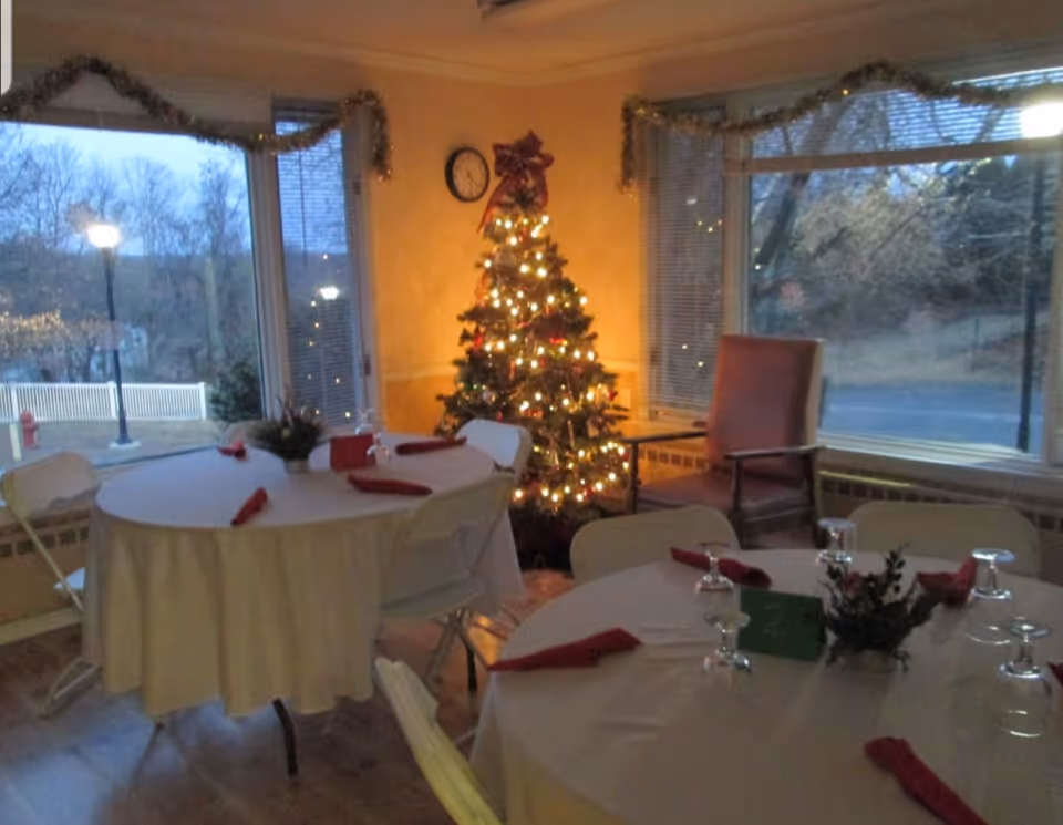 A cozy dining area decorated for Christmas with a lit Christmas tree in the corner, round tables covered with white tablecloths, red napkins, upside-down wine glasses, and small floral centerpieces. Large windows show an outdoor view with trees and street lamps, and garlands hang along the top of the windows.