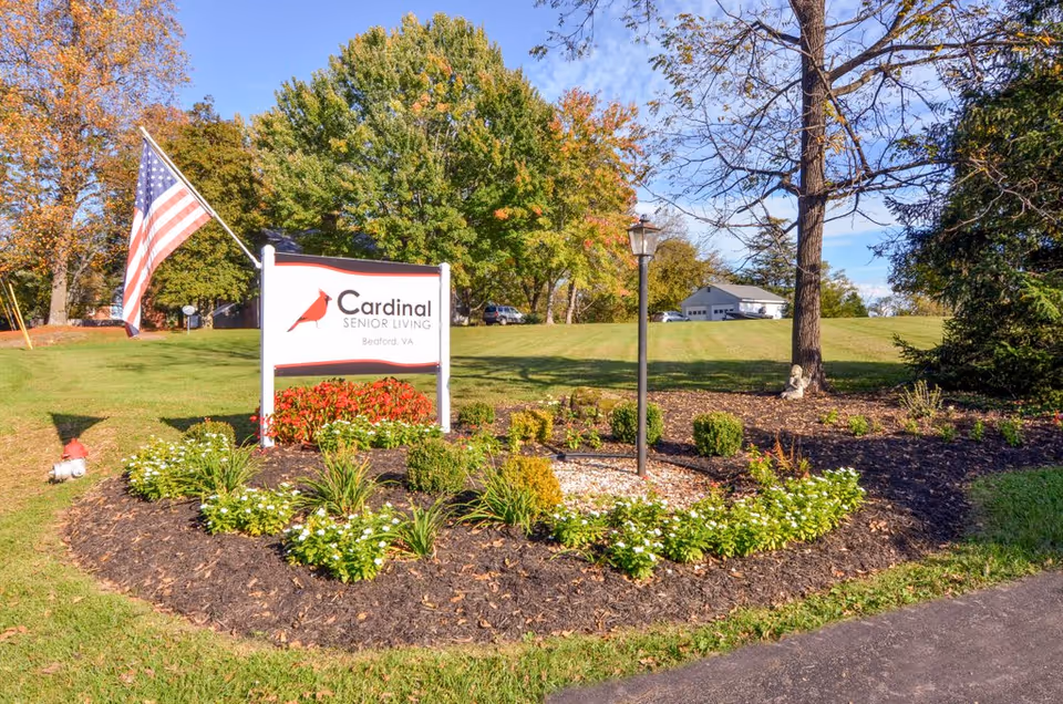 Outdoor landscaped area with a sign for Cardinal Senior Living Bedford, VA, surrounded by flowers and greenery, an American flag on a pole, and a lamp post, with trees and a building in the background under a clear blue sky.