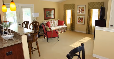 Bright living room with a floral sofa, red armchair, TV, and a kitchen island with bar stools.