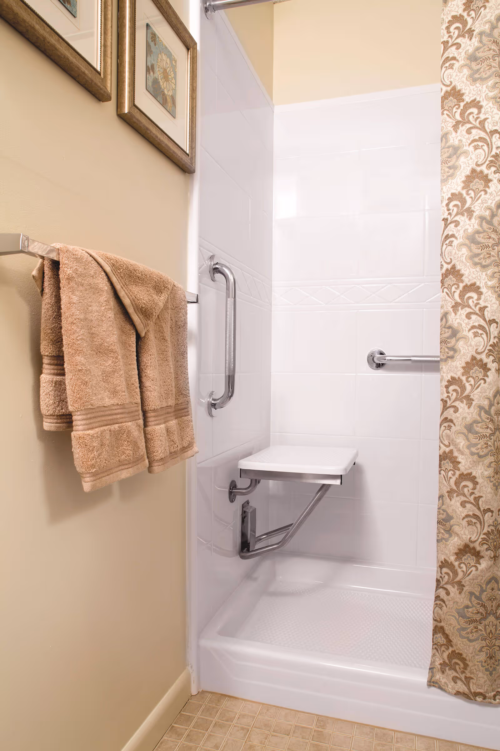 Shower stall with a fold-down seat, grab bars, a patterned shower curtain, and towels hanging on a nearby rack.