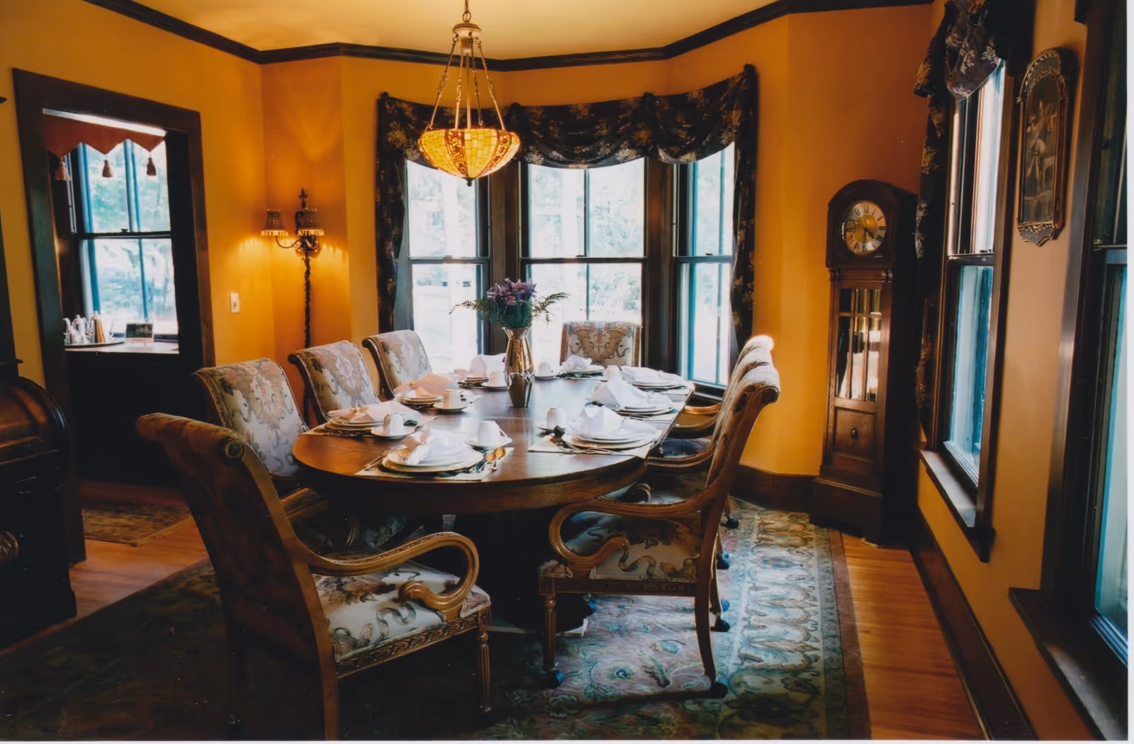 A warmly lit dining room with a large oval wooden table set for eight people. The table is adorned with white napkins, plates, cups, and a vase with purple flowers in the center. The room features yellow walls, large windows with dark floral curtains, a hanging chandelier, a grandfather clock, and a patterned rug beneath the table and chairs.