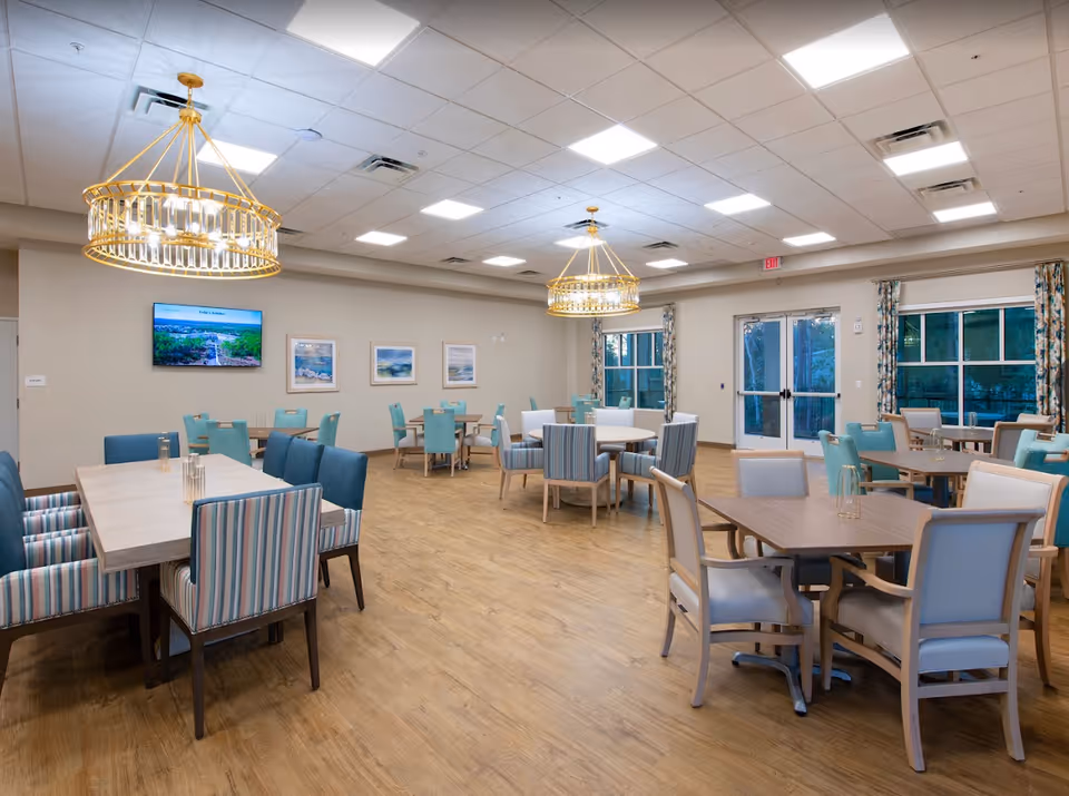 A spacious dining room with multiple tables and chairs arranged neatly. The room features wooden flooring, large windows with patterned curtains, and two large modern chandeliers hanging from the ceiling. A flat-screen TV is mounted on the wall displaying an image of a landscape, and framed artwork is hung on the wall beneath it. The room is well-lit with ceiling lights and has double glass doors leading outside.