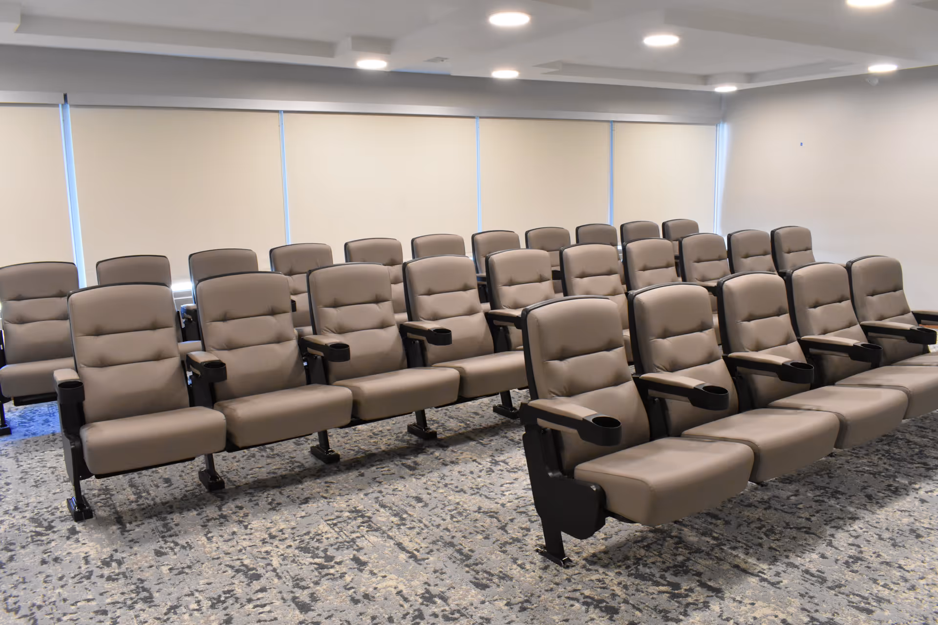 A room with multiple rows of beige cushioned chairs with armrests and cup holders, arranged in a theater or presentation style. The room has a carpeted floor with a patterned design and windows covered with beige blinds.