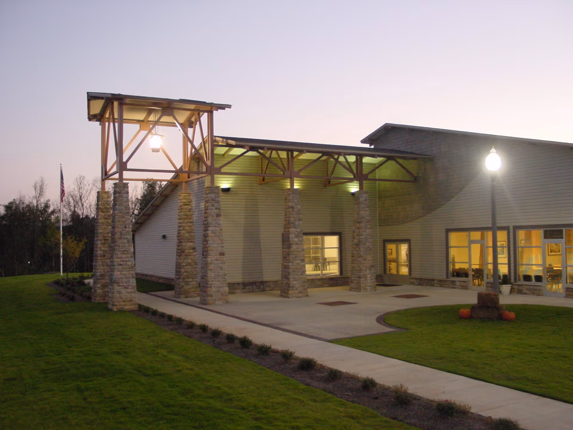 Exterior view of Oaks on Parkwood facility at dusk, showing a covered entrance supported by stone pillars, a well-maintained lawn, a sidewalk, an American flag on a flagpole, and a lit streetlamp near the building entrance with windows revealing interior lighting.
