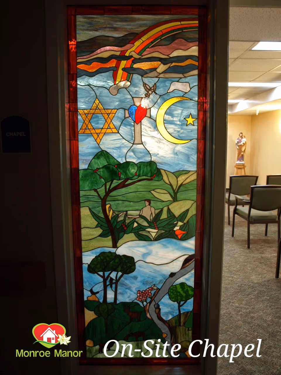 A tall stained-glass window with religious symbols and a pastoral scene set in a doorway opening into a small chapel with chairs.