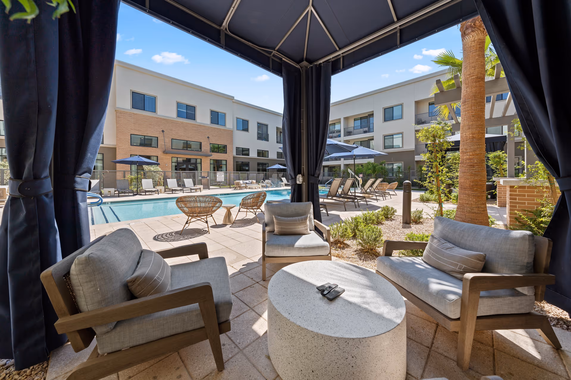 Covered cabana seating area with armchairs and a round table overlooking a pool and surrounding three-story building.