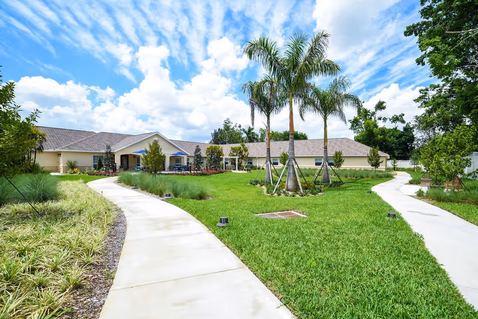 Outdoor view of a senior living facility with a curved concrete pathway leading through a well-maintained green lawn with palm trees and landscaped plants. The building is single-story with a beige exterior and a gray roof under a partly cloudy blue sky.