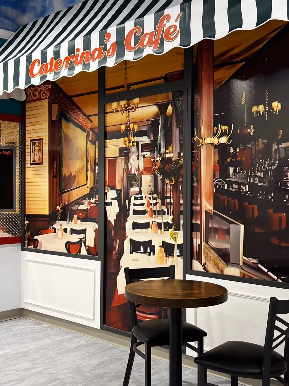 A small seating area with a round wooden table and two black chairs in front of a wall mural depicting the interior of a cozy café named Caterina's Café with tables covered in white tablecloths and warm lighting.