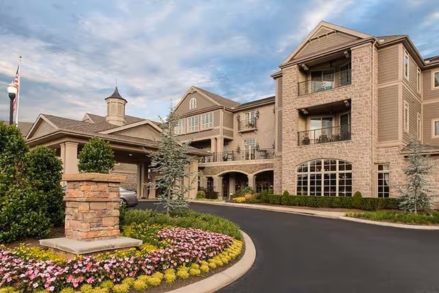 Exterior view of a senior living facility with a multi-story building featuring balconies and large windows. The entrance has a covered driveway with stone pillars and landscaped flower beds with pink and yellow flowers. The sky is partly cloudy.