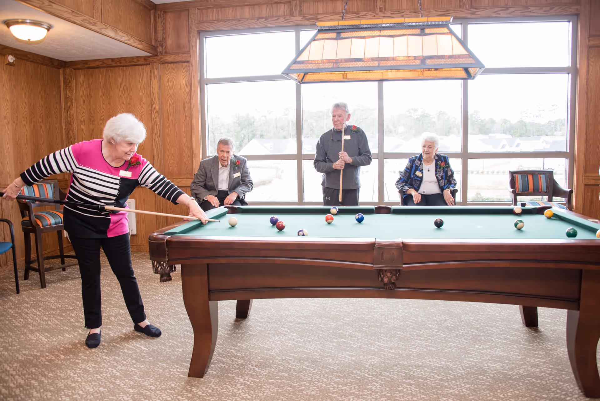 Four elderly people in a wood-paneled room with large windows playing pool. One woman is taking a shot while two people sit on chairs and one man stands holding a cue stick. The room has a carpeted floor and a large hanging light fixture above the pool table.