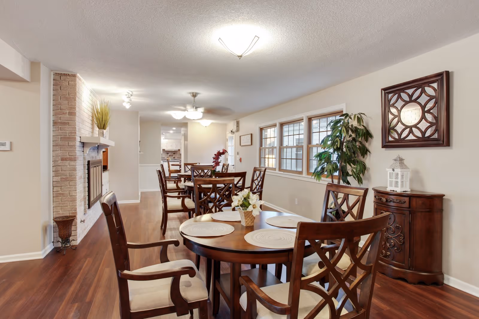 A bright dining room with wooden floors and multiple round wooden tables surrounded by cushioned chairs. The room features a brick fireplace on the left wall, a large window on the right side, a decorative mirror, a potted plant, and a small wooden cabinet. The ceiling has modern light fixtures and a ceiling fan.