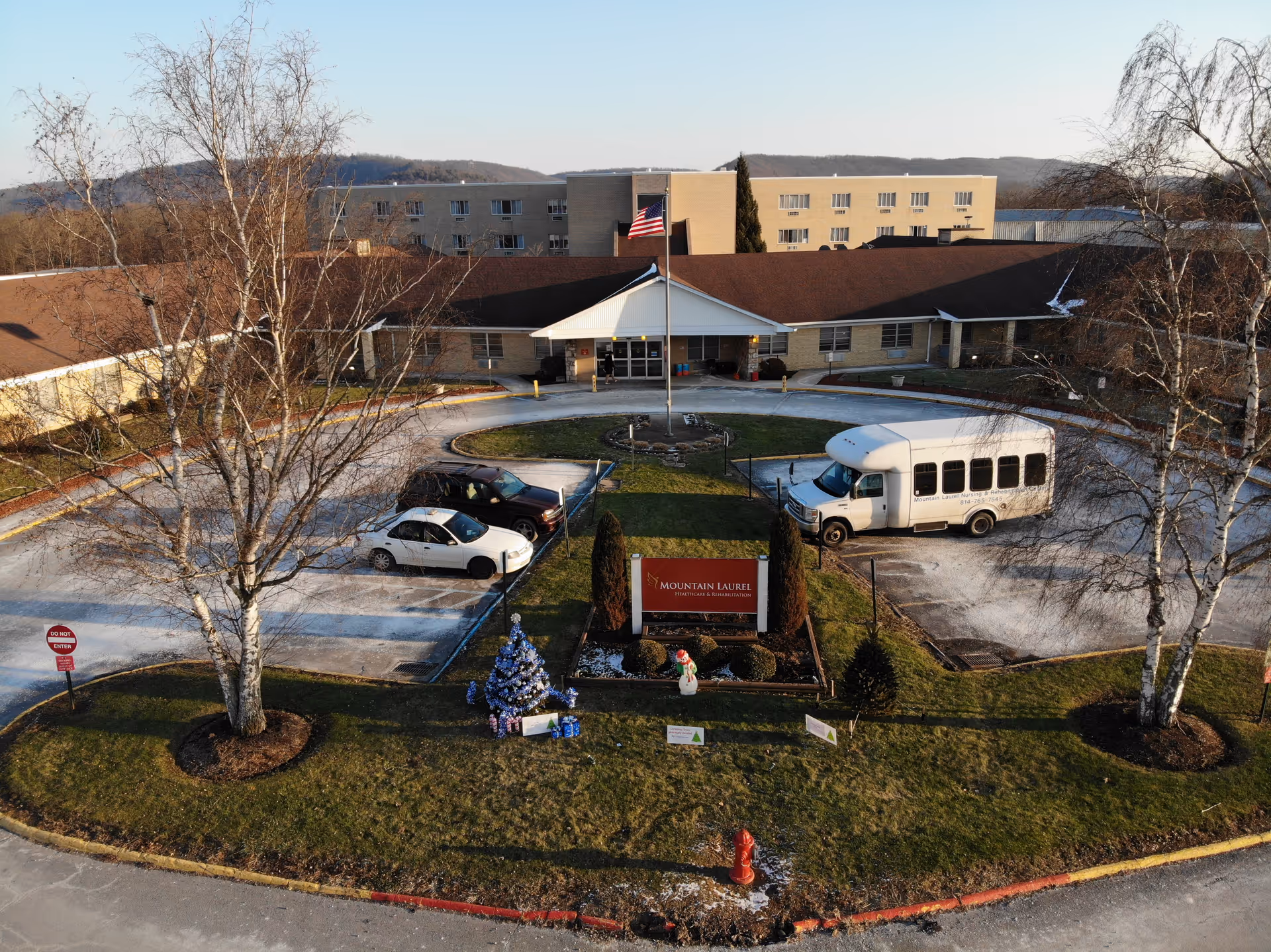 Front exterior view of Mountain Laurel Healthcare and Rehabilitation Center showing a circular driveway with parked vehicles, a flagpole with an American flag, leafless trees, a decorated Christmas tree, and a snowman figure on the lawn.
