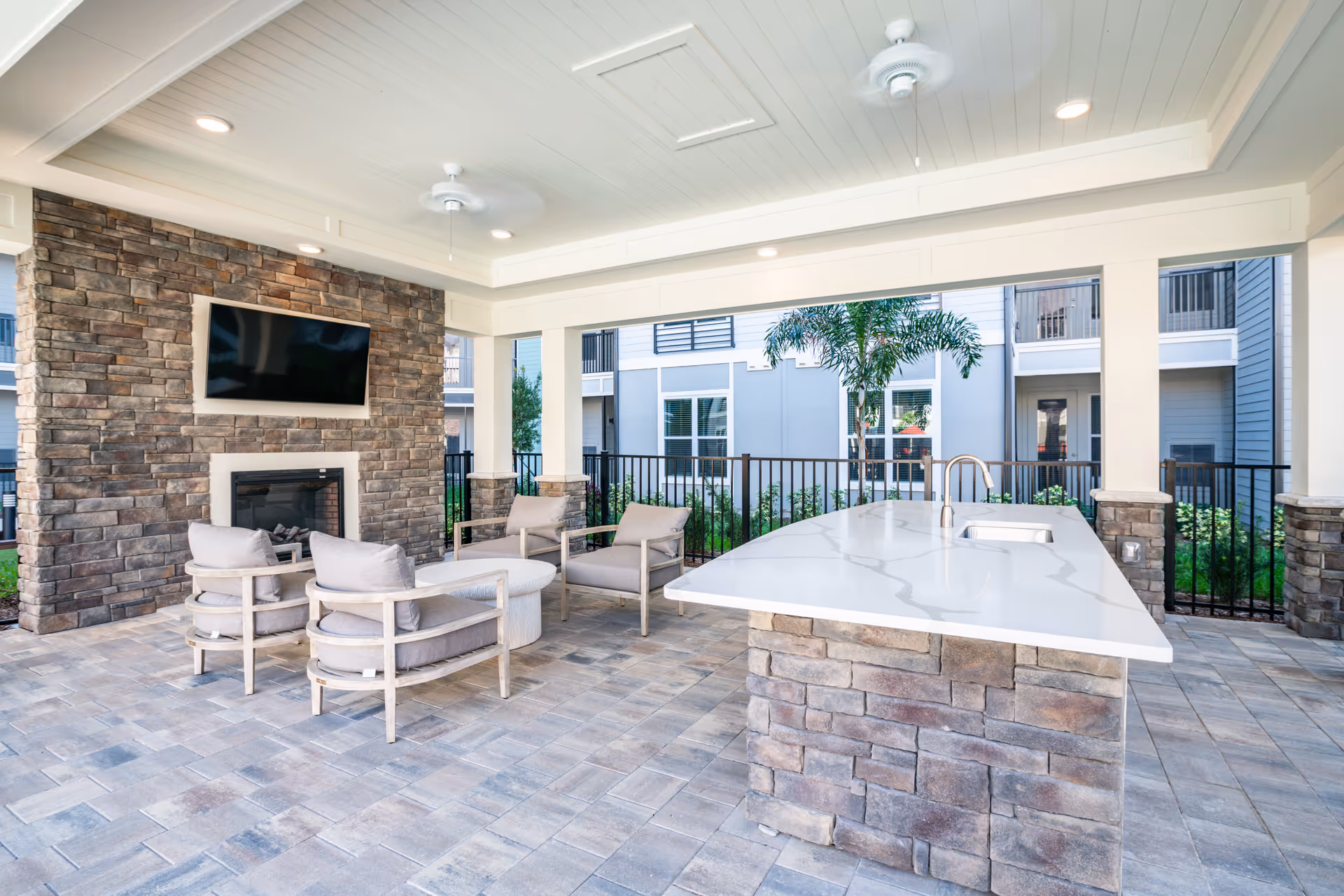 Covered outdoor lounge with stone fireplace, wall-mounted TV, cushioned seating and a large stone island countertop.