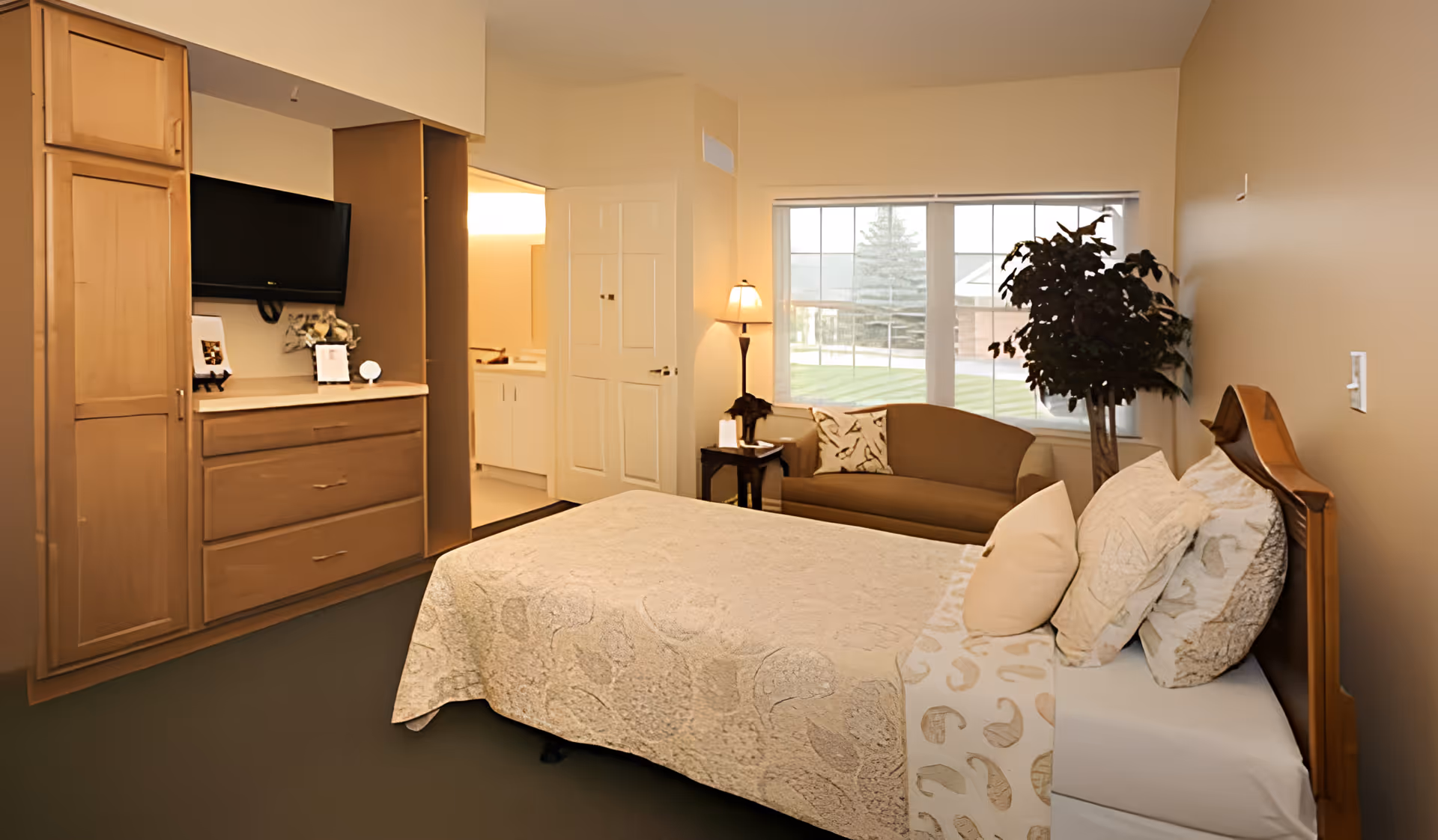 A cozy bedroom in Hartsfield Village featuring a bed with multiple pillows and a patterned quilt, a wooden headboard, a small brown sofa with a decorative pillow, a floor lamp, a large window with a view of the outdoors, and built-in wooden cabinetry with a mounted flat-screen TV. An open door reveals a glimpse of a bathroom area.