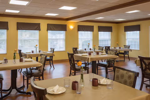A bright dining room with multiple tables covered in beige tablecloths, each set with plates, glasses, napkins, and small vases holding orange flowers. The room has large windows with brown valances, yellow walls, and wooden flooring.
