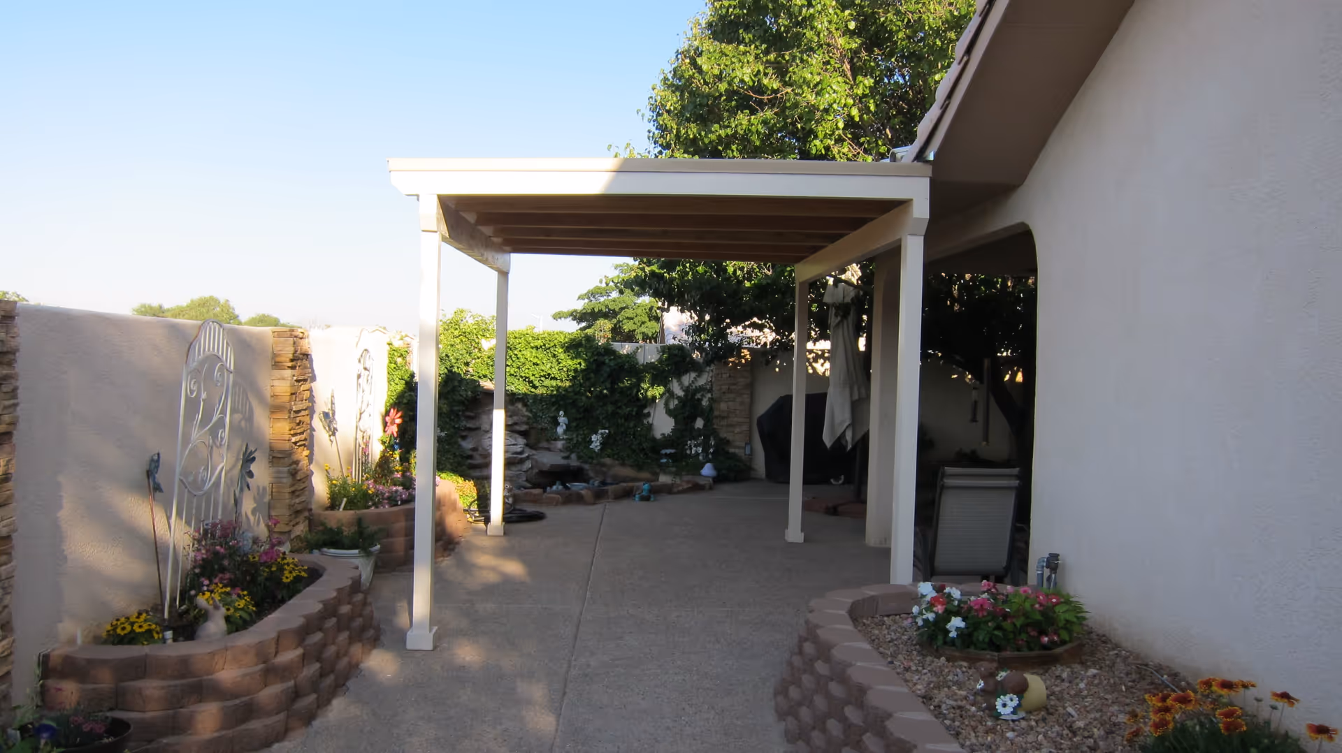 Outdoor patio area with a pergola providing shade, surrounded by garden beds with flowers and decorative elements, adjacent to a beige stucco building wall.