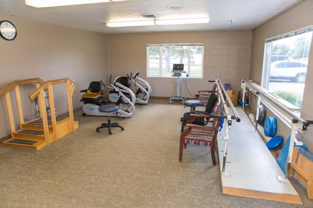 A rehabilitation room with exercise equipment including stationary bikes, parallel bars for walking practice, chairs, and a small wooden staircase for physical therapy. The room has beige carpet, beige walls, a clock on the wall, and windows letting in natural light.