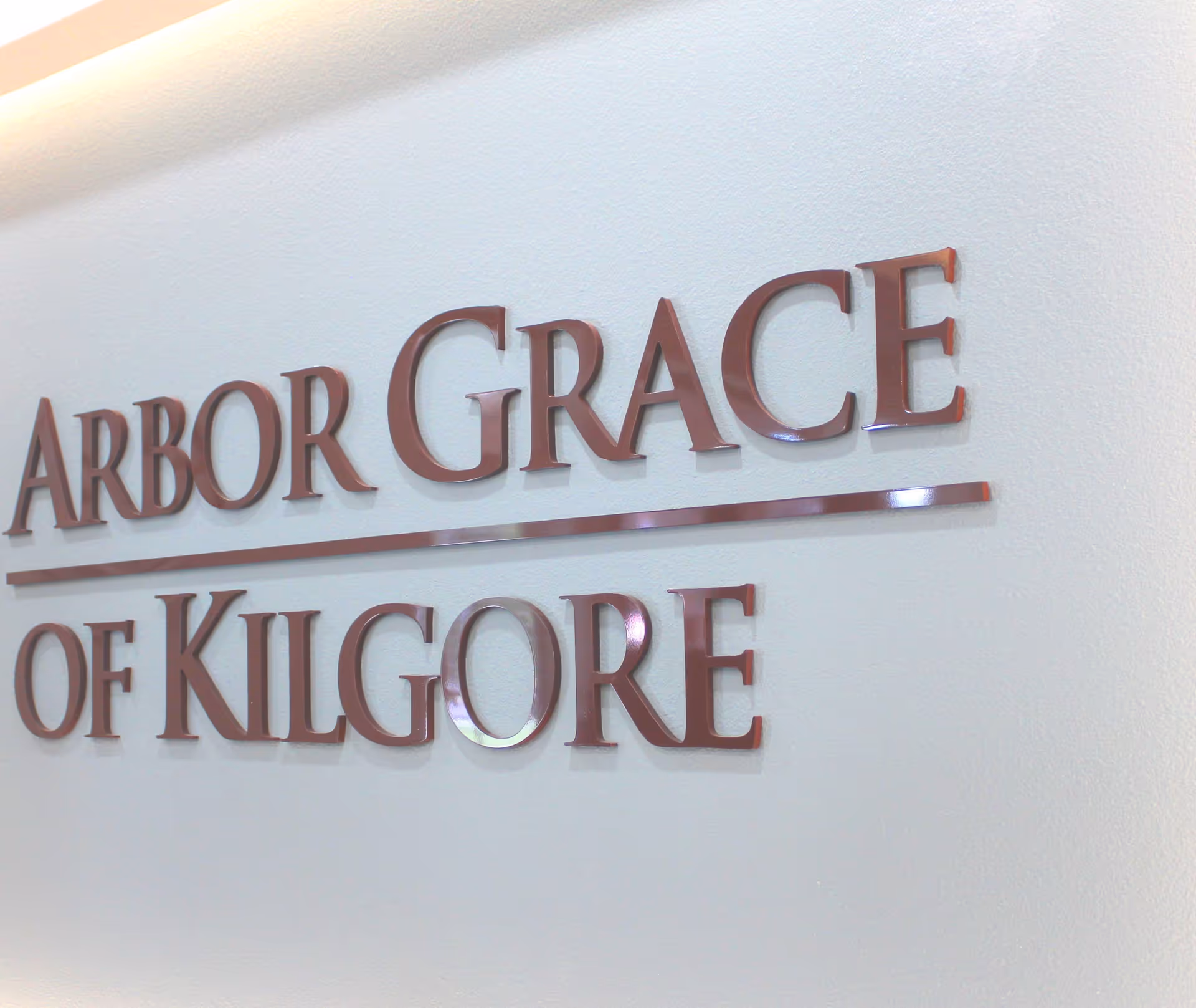 Close-up view of a wall sign with raised metallic letters spelling 'ARBOR GRACE of KILGORE' on a light-colored textured wall.