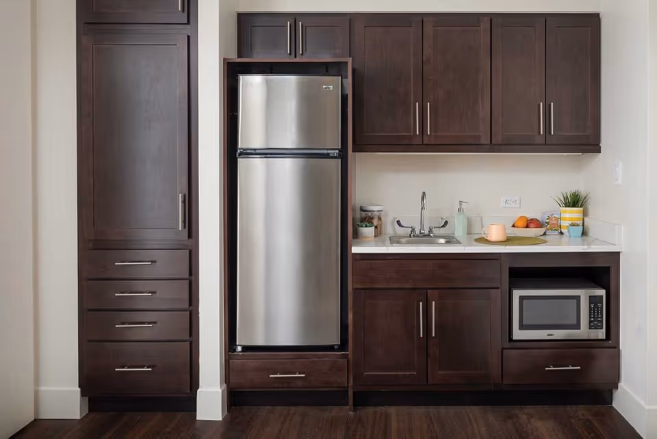 Small kitchenette with dark wood cabinets, a stainless steel refrigerator, sink, microwave, and countertop accessories.
