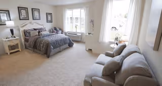 A spacious and well-lit bedroom featuring a large bed with gray bedding, a nightstand with a lamp, framed artwork on the wall, a window with white curtains, a small table with flowers, and a gray sofa with cushions.