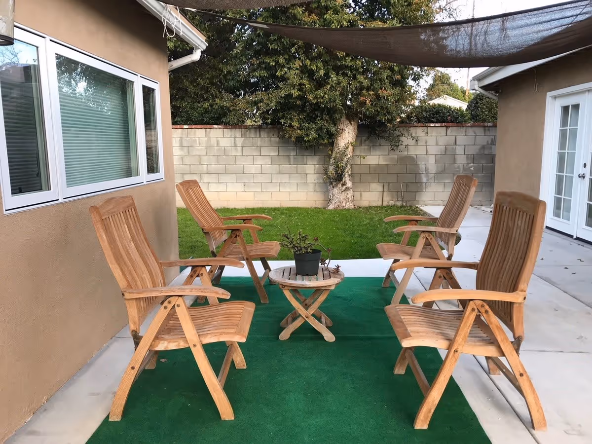 Outdoor patio with six wooden chairs arranged around a small round table on a green outdoor rug beside a stucco building and a cinderblock wall.