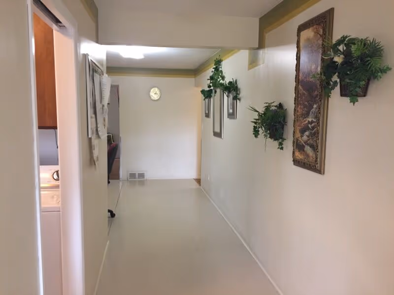 A long, narrow hallway with beige walls and a light-colored floor. The right wall is decorated with framed artwork and several green potted plants mounted on the wall. At the end of the hallway, there is a round wall clock. On the left side near the foreground, a doorway leads to a laundry area with a washing machine visible.