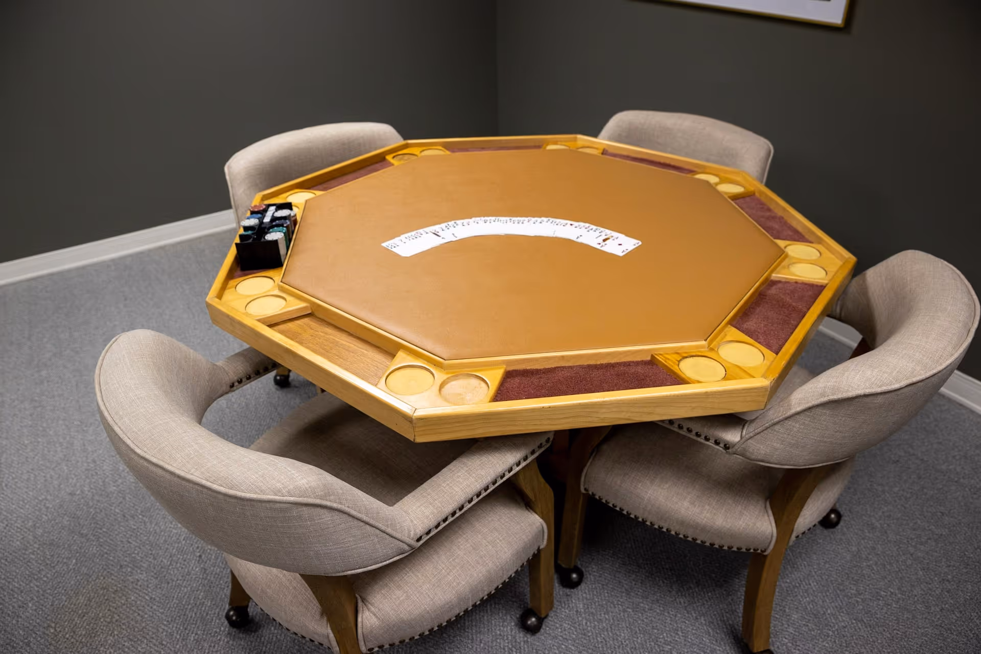 Octagonal wooden card table with playing cards fanned in the center surrounded by four upholstered chairs in a carpeted room.