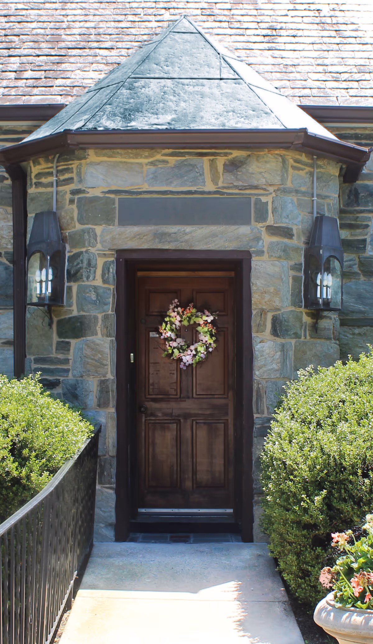 A stone building entrance with a dark wooden door decorated with a floral wreath. The entrance is flanked by two wall-mounted lantern-style lights and surrounded by green bushes. A paved pathway leads up to the door.