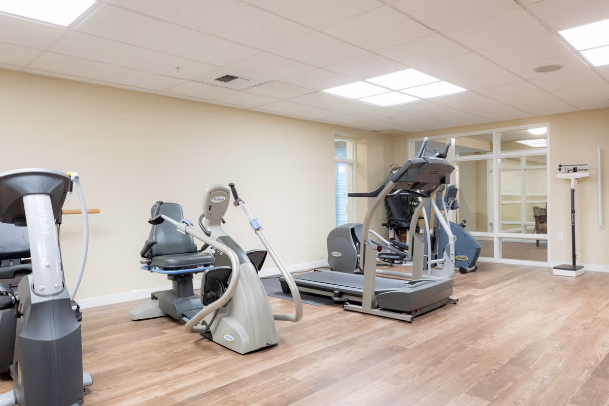 A fitness room with exercise equipment including a treadmill, stationary bikes, and an elliptical machine. The room has light-colored walls, wood flooring, and a large window with glass panels. A scale is visible in the corner.