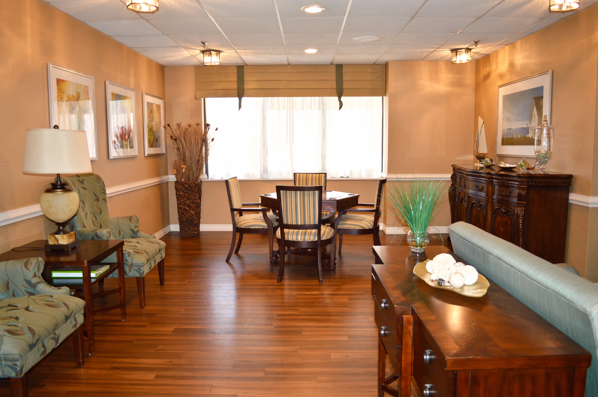 A warmly lit interior room with wooden flooring featuring a small dining table with four striped upholstered chairs near a window with beige curtains. To the left, there are two patterned armchairs with a side table and lamp between them. On the right side, there is a wooden sideboard with decorative items and a green cushioned bench in front of it. The walls are adorned with framed artwork and a tall vase with dried branches is placed near the window.