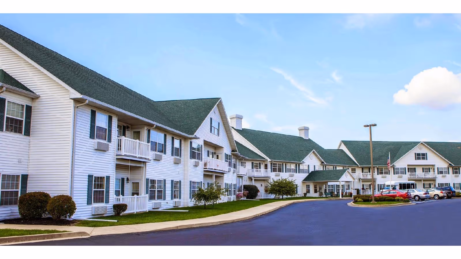 Large two-story white senior living building with green roofs, balconies, a driveway and parked cars under a blue sky.