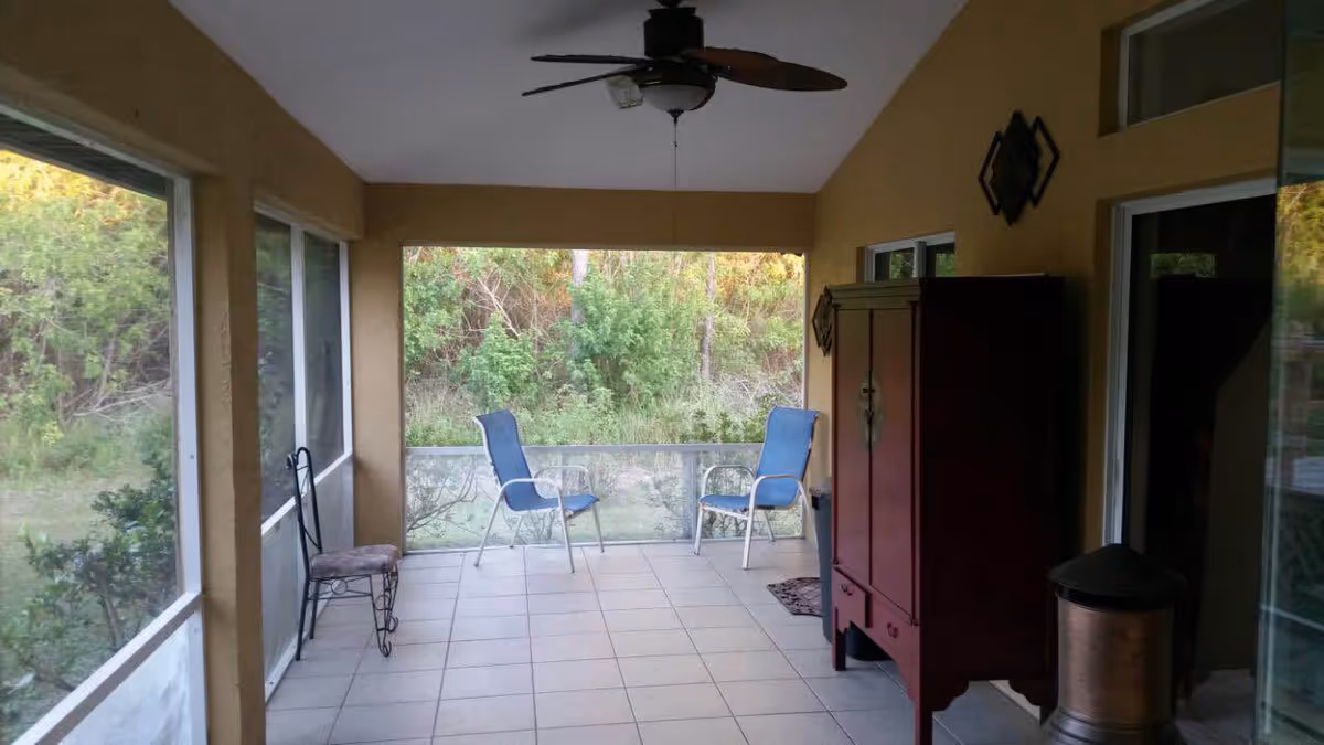 A screened-in porch with tiled flooring, two blue chairs facing outward, a small decorative chair on the left, a ceiling fan above, and a wooden cabinet on the right side. The porch overlooks a green, wooded area.