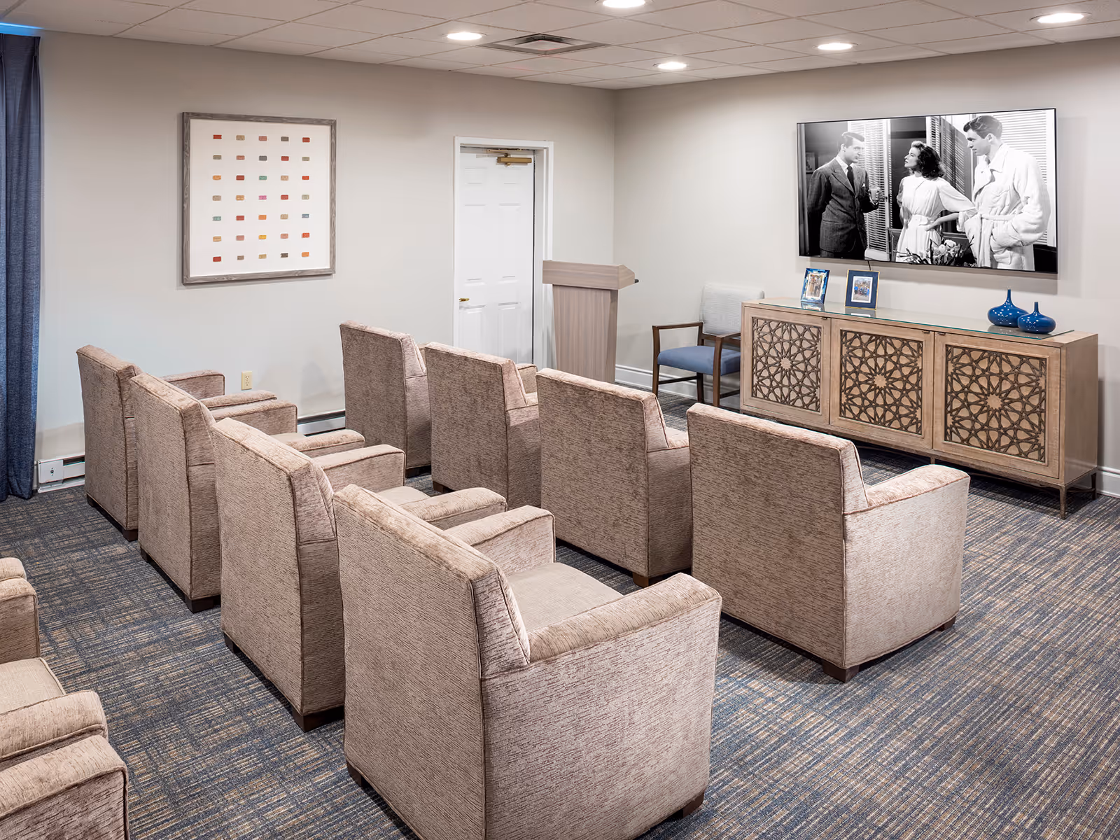 A small theater or media room with two rows of beige upholstered armchairs facing a large flat-screen TV mounted on the wall. Below the TV is a wooden cabinet with decorative carved doors, two framed photos, and two blue vases. There is a podium near a white door, and a framed abstract artwork on the adjacent wall. The room has a carpeted floor and a drop ceiling with recessed lighting.