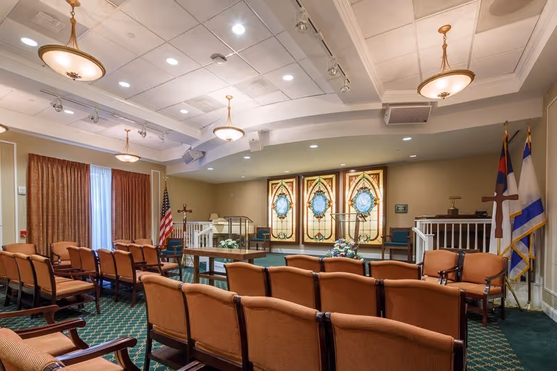 Interior view of a chapel or worship room with rows of brown cushioned chairs facing a stage area with stained glass windows, flags, and a wooden cross. The room has a green carpet with a patterned design, beige walls, and ceiling lights.
