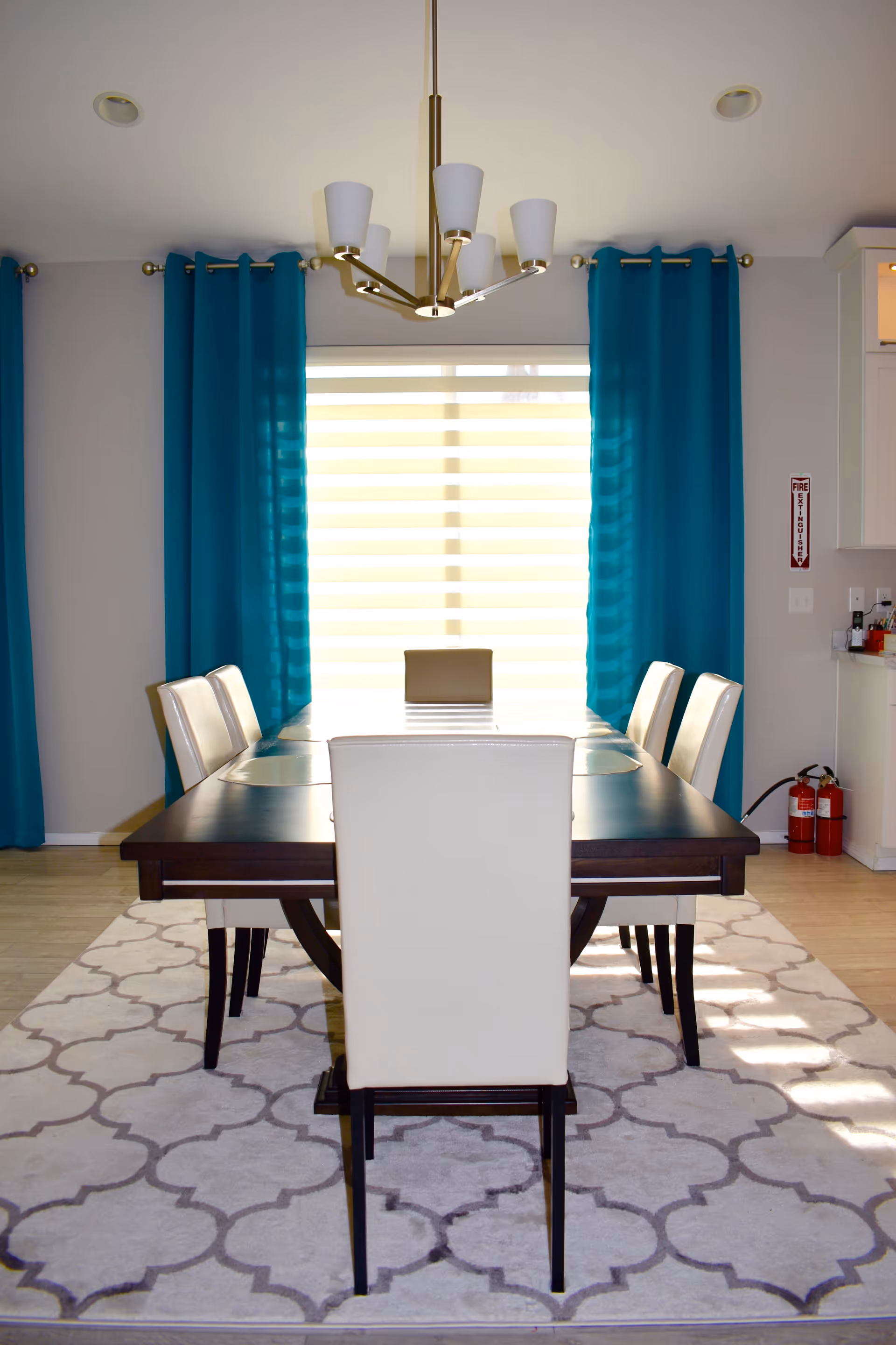 A dining room with a dark wooden table surrounded by six cream-colored chairs. The room has a patterned light gray and white rug underneath the table. Bright natural light comes through a large window covered with white blinds and flanked by teal curtains. A modern chandelier with five lights hangs above the table. Two fire extinguishers are visible near the kitchen area on the right side of the image.