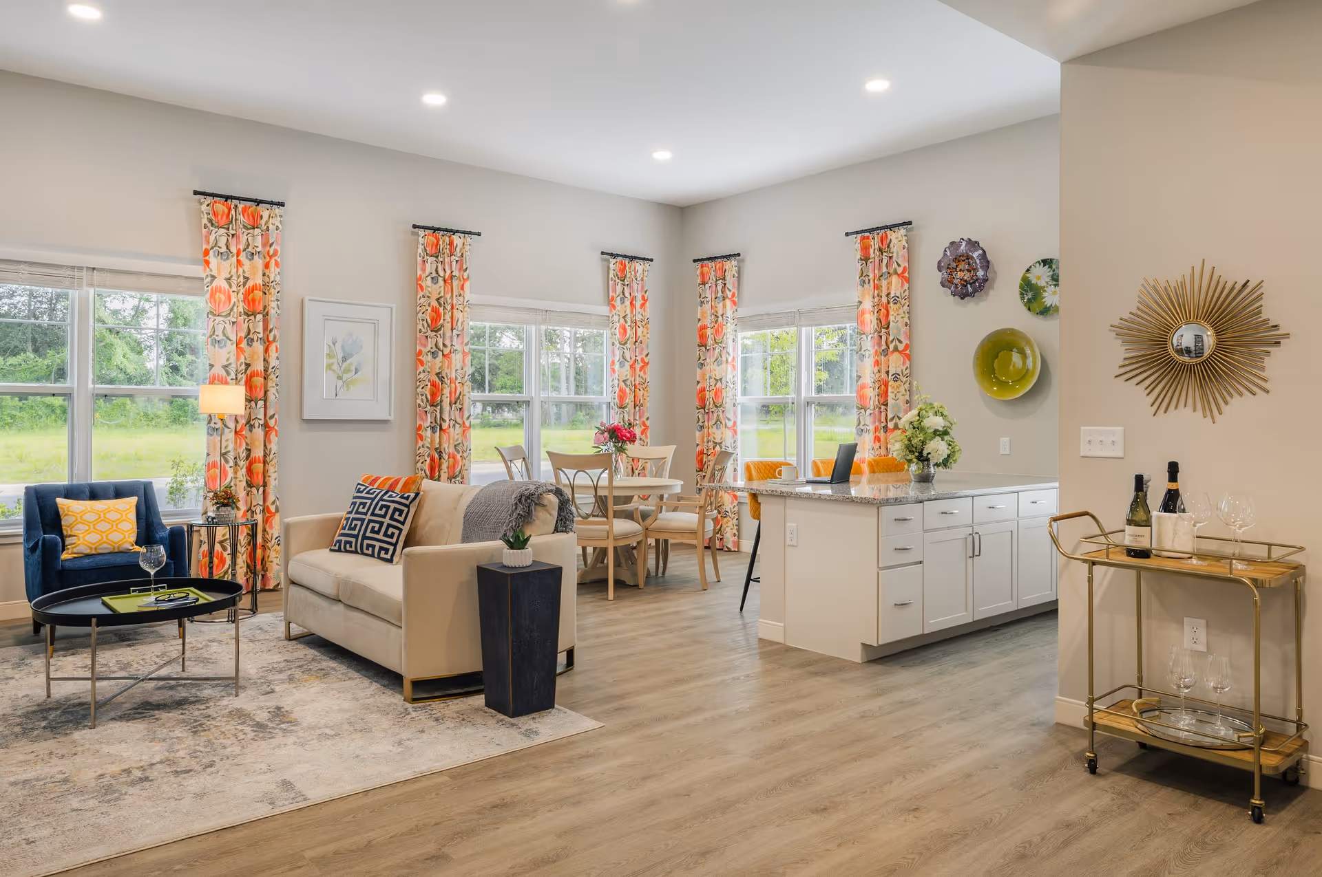 Open-concept living area with a sofa and armchair, dining table, kitchen island, and large windows with floral curtains.