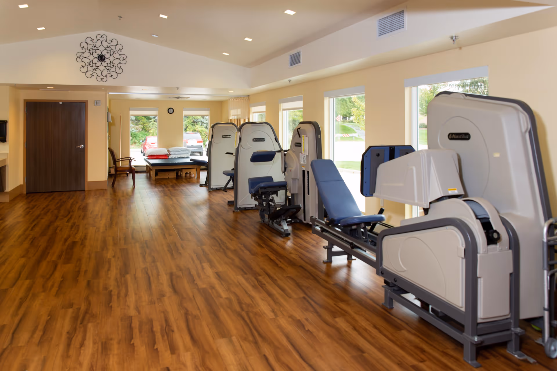 A bright exercise room with wooden flooring and several pieces of Nautilus fitness equipment arranged along the right side. Large windows on the right wall let in natural light and offer a view of greenery outside. At the far end of the room, there is a treatment table with folded towels and pillows, and a wooden chair next to a closed wooden door. The ceiling is vaulted with recessed lighting and a decorative wall clock is mounted above the door.