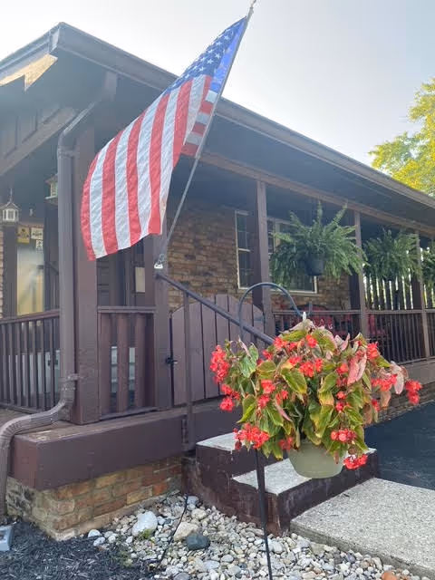 Front porch of a brown building with an American flag and hanging red flowers by the entrance.