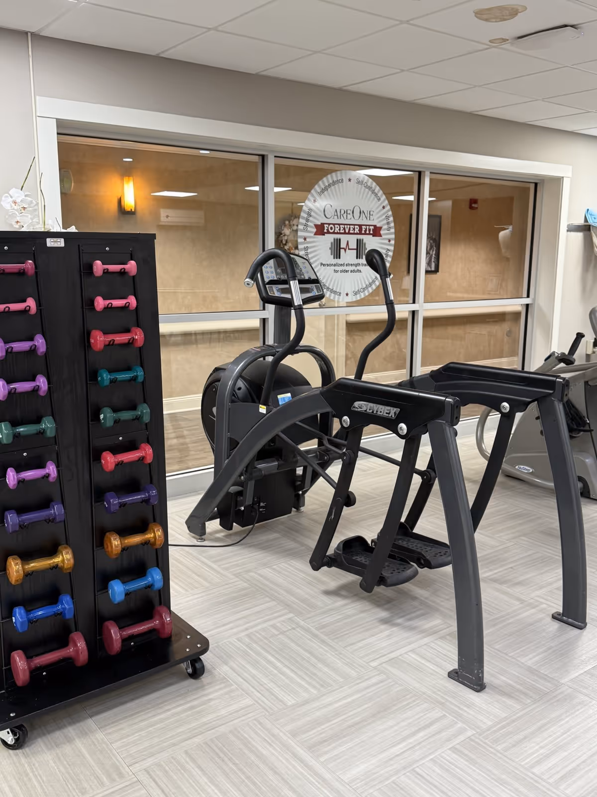 Indoor fitness room with exercise equipment including a Cybex machine and a rack of colorful dumbbells. A large window in the background displays a sign that reads 'CareOne Forever Fit Personalized strength training for older adults.'