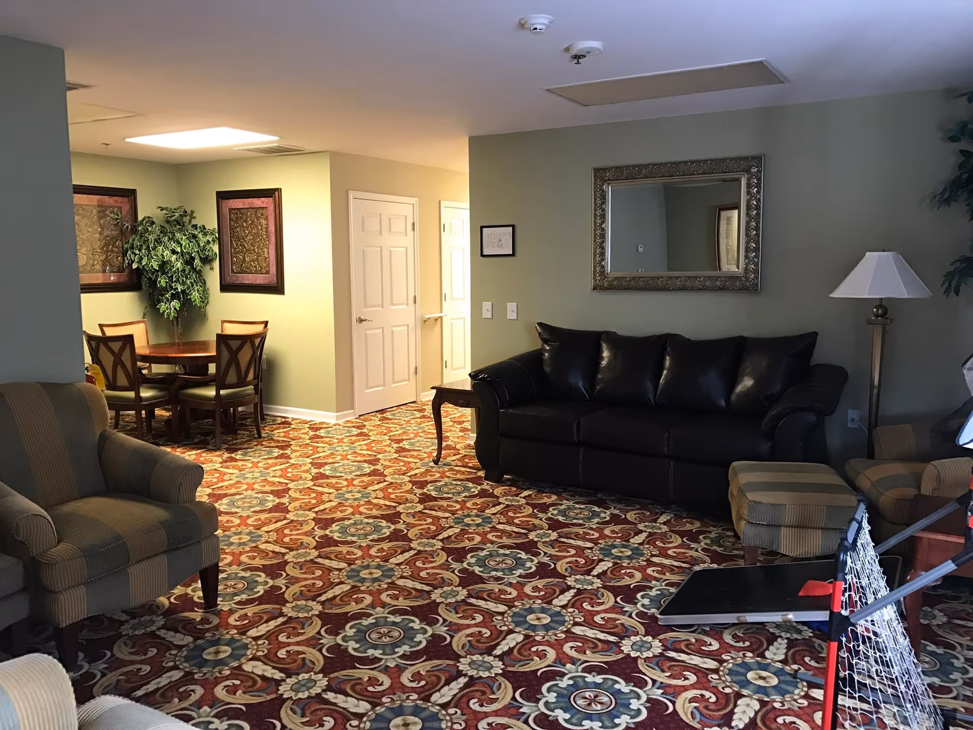 Comfortable assisted-living common room with a leather sofa, armchairs, a small dining table, and a patterned carpet.