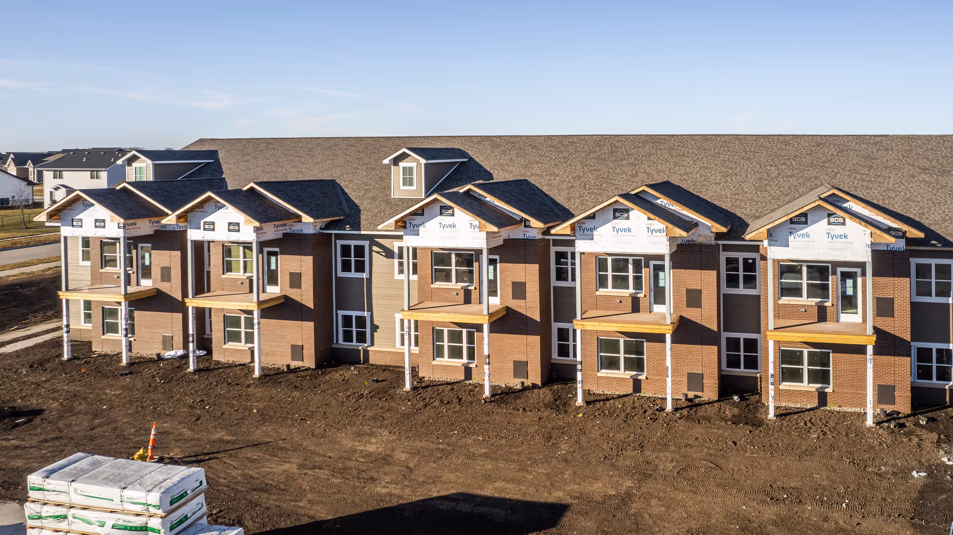 A large two-story building under construction with multiple small balconies and windows. The exterior walls are partially covered with brick and siding, and some areas are wrapped with Tyvek building wrap. The ground in front of the building is bare soil with construction materials stacked nearby.