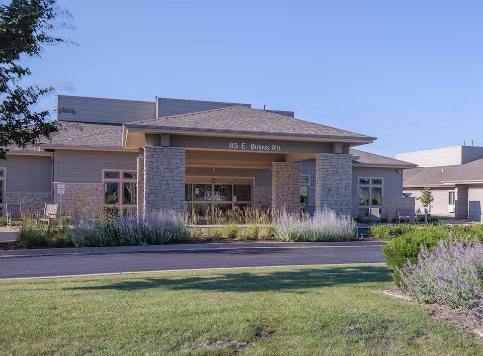 Front entrance of a single-story senior living building with stone columns, a covered portico reading '85 E. Burns Rd.', and landscaped lawn and shrubs in front.