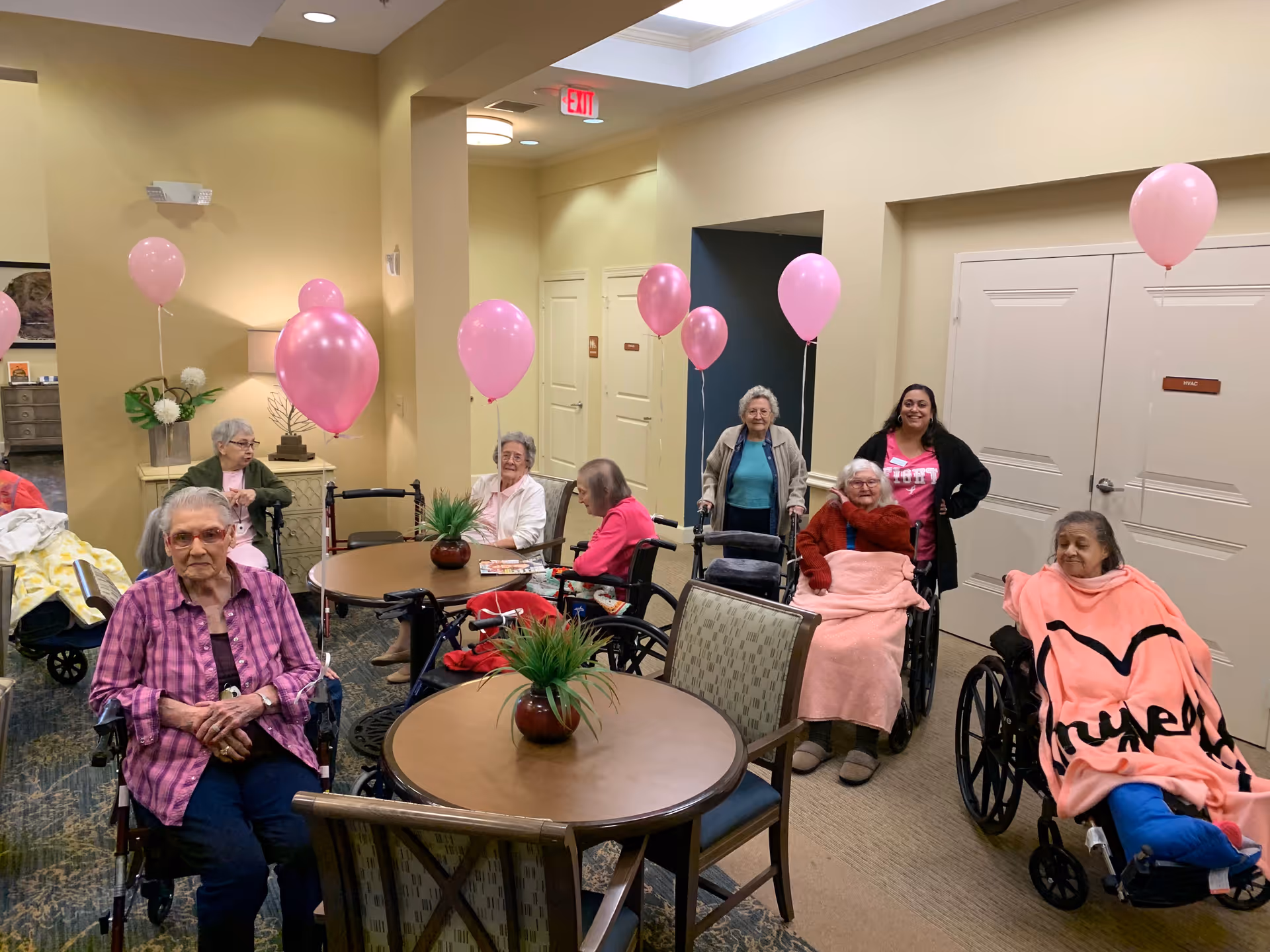 Residents seated in a senior living common room around tables and in wheelchairs with pink balloons.