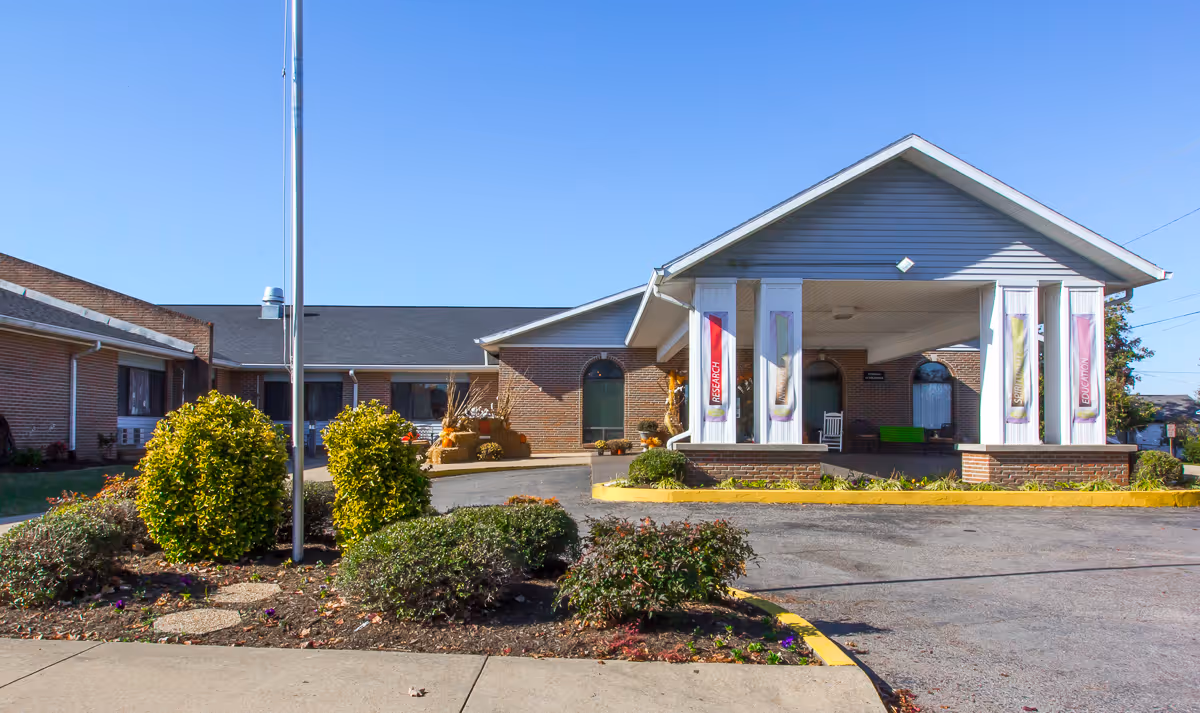 Front exterior view of Signature HealthCARE of Ridgely Rehab & Wellness Center building with a covered entrance, brick walls, and landscaped bushes under a clear blue sky.