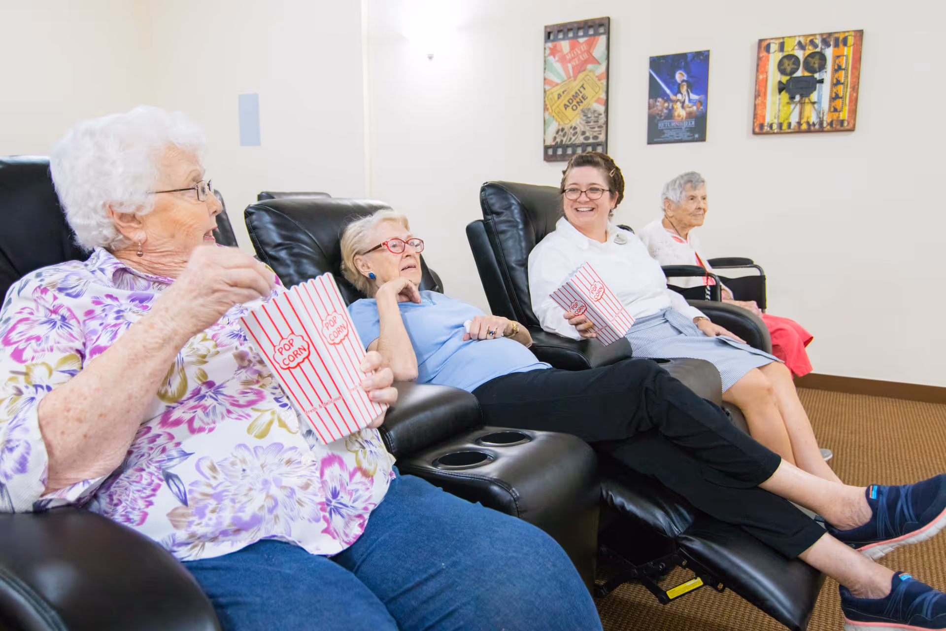 Four elderly women sitting in black reclining chairs in a room with movie-themed posters on the wall. Three of the women are holding striped popcorn containers and appear to be enjoying a movie or social activity together.