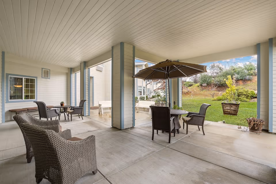 Covered outdoor patio area with wicker chairs, tables, and an umbrella, overlooking a grassy garden with shrubs and trees.