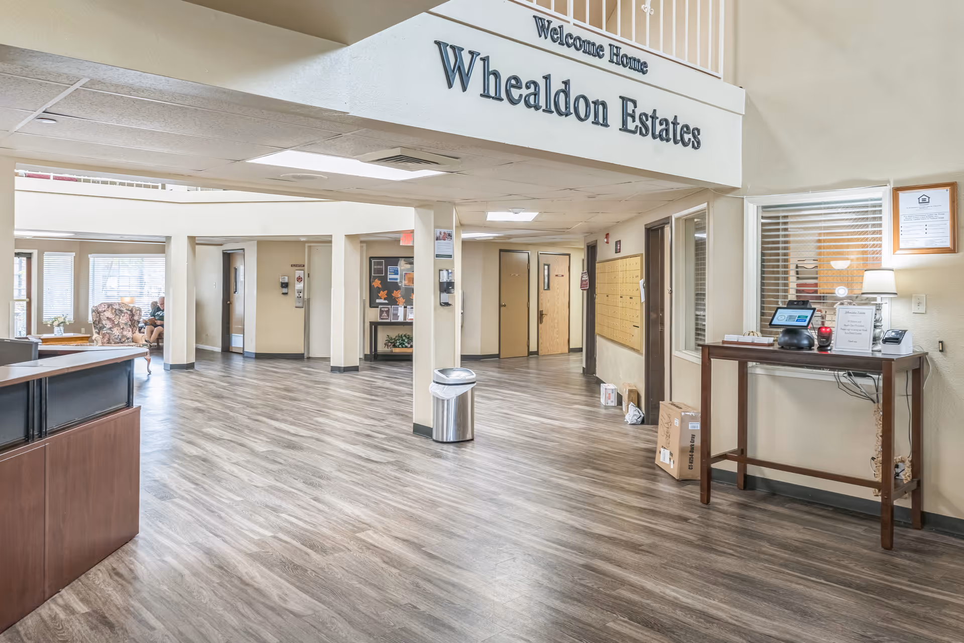 Interior view of the entrance lobby of Whealdon Estates by Barclay House, featuring a reception desk on the left, a wooden table with a lamp and electronic device on the right, mailboxes on the wall, and a seating area with chairs and windows in the background. The floor has wood-like vinyl flooring and the ceiling has recessed lighting. A sign above reads 'Welcome Home Whealdon Estates'.