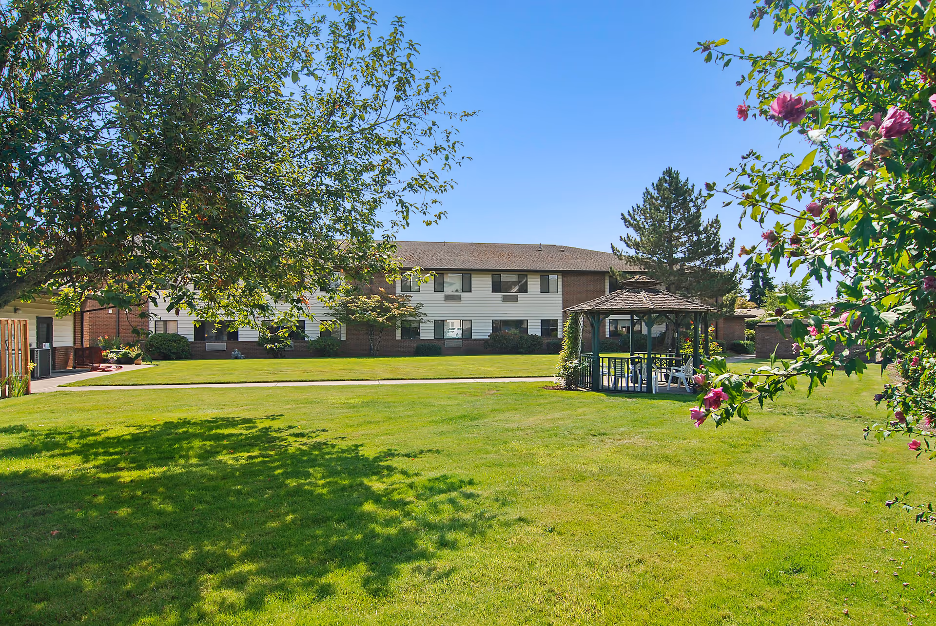 A sunny outdoor garden area with a well-maintained green lawn, a wooden gazebo with chairs underneath, surrounded by trees and flowering bushes. In the background, there is a two-story building with white siding and brick accents under a clear blue sky.