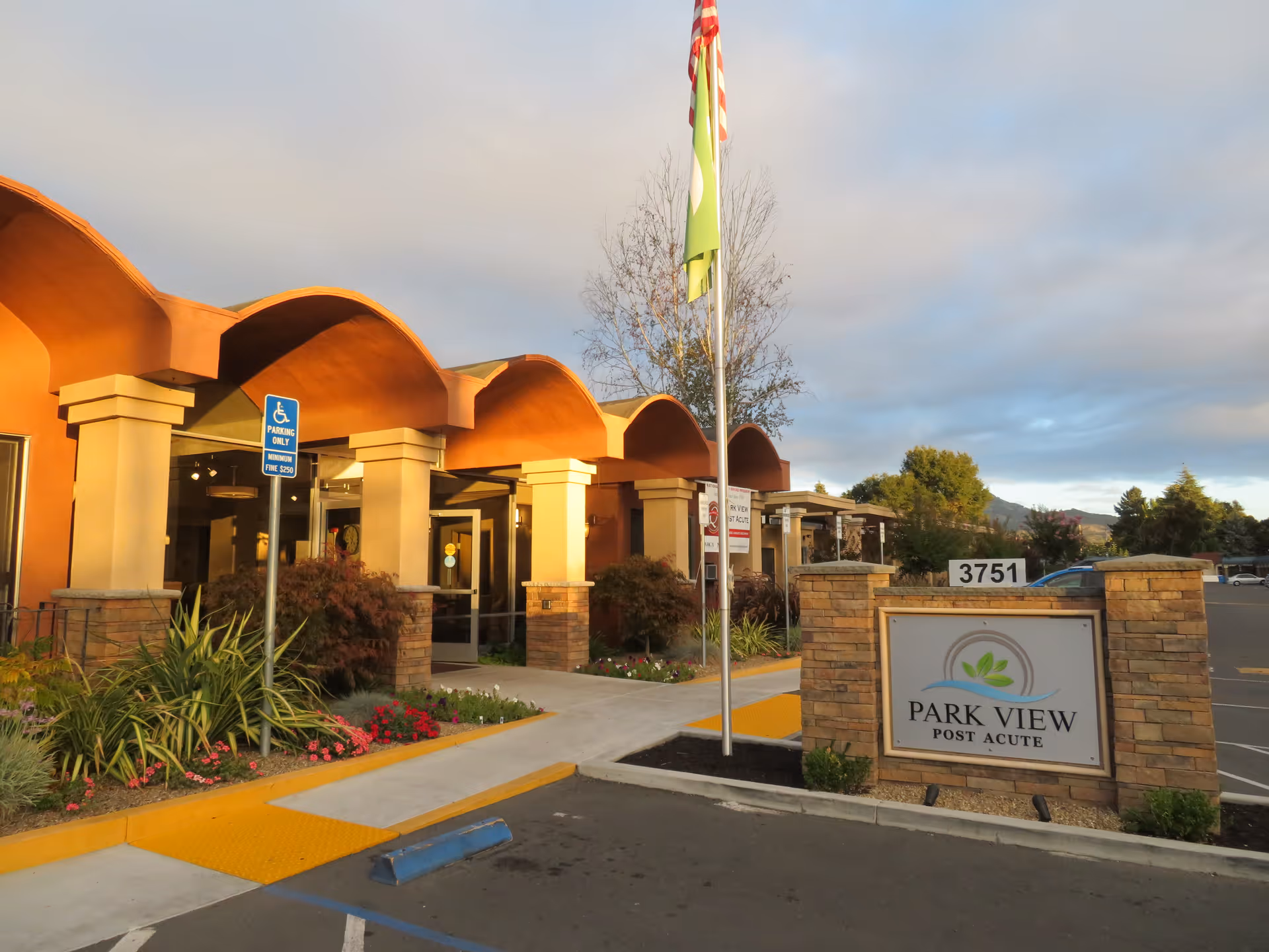 Exterior view of Park View Post Acute facility showing the entrance with a covered walkway, landscaping with plants and flowers, a flagpole with flags, and a sign displaying the facility name and address number 3751.